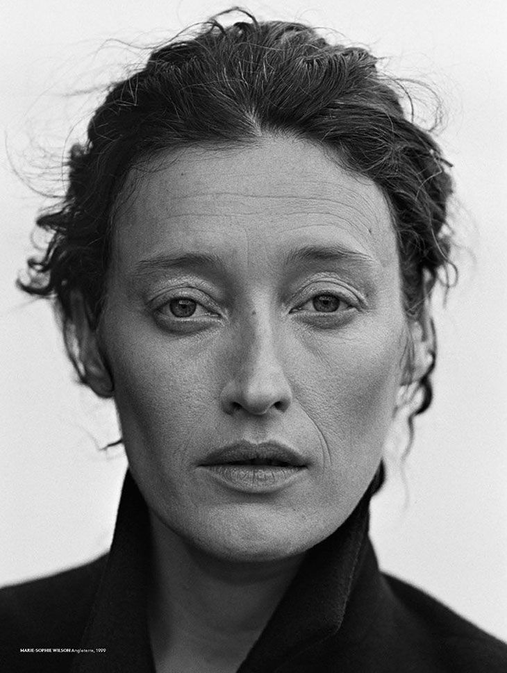 a black and white portrait of a mature woman with her hair pulled back, revealing her face and neck. the image has low contrast, emphasizing her wrinkles and natural features. she has a serious expression and is wearing dark clothing with a high collar. the photograph was taken by marie sophie wilson in 1999.