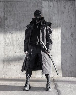 a man is wearing a long black coat, black pants, black boots, a black mask, and a black hat. he is holding a katana in his right hand. he is standing in front of a concrete wall. he is looking at the camera.