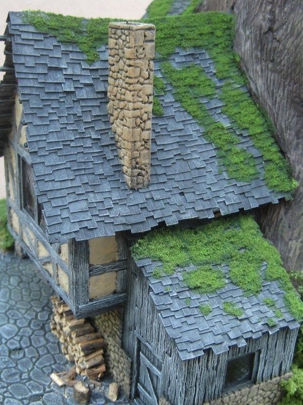 an image of a miniature rustic house with a stone chimney, a shingled roof covered in moss, and wooden walls. the house is part of a diorama or model scene. there's a stack of wood next to the house.