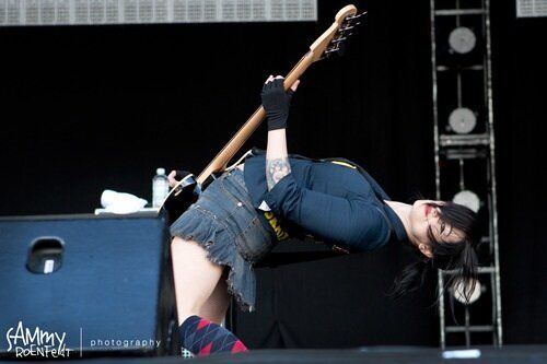 a woman with long dark hair and a tattoo on her arm is playing a bass guitar. she is bending over with her head tilted back and her eyes are looking towards the camera. she is wearing jeans and a dark shirt. she is performing on stage at a concert.