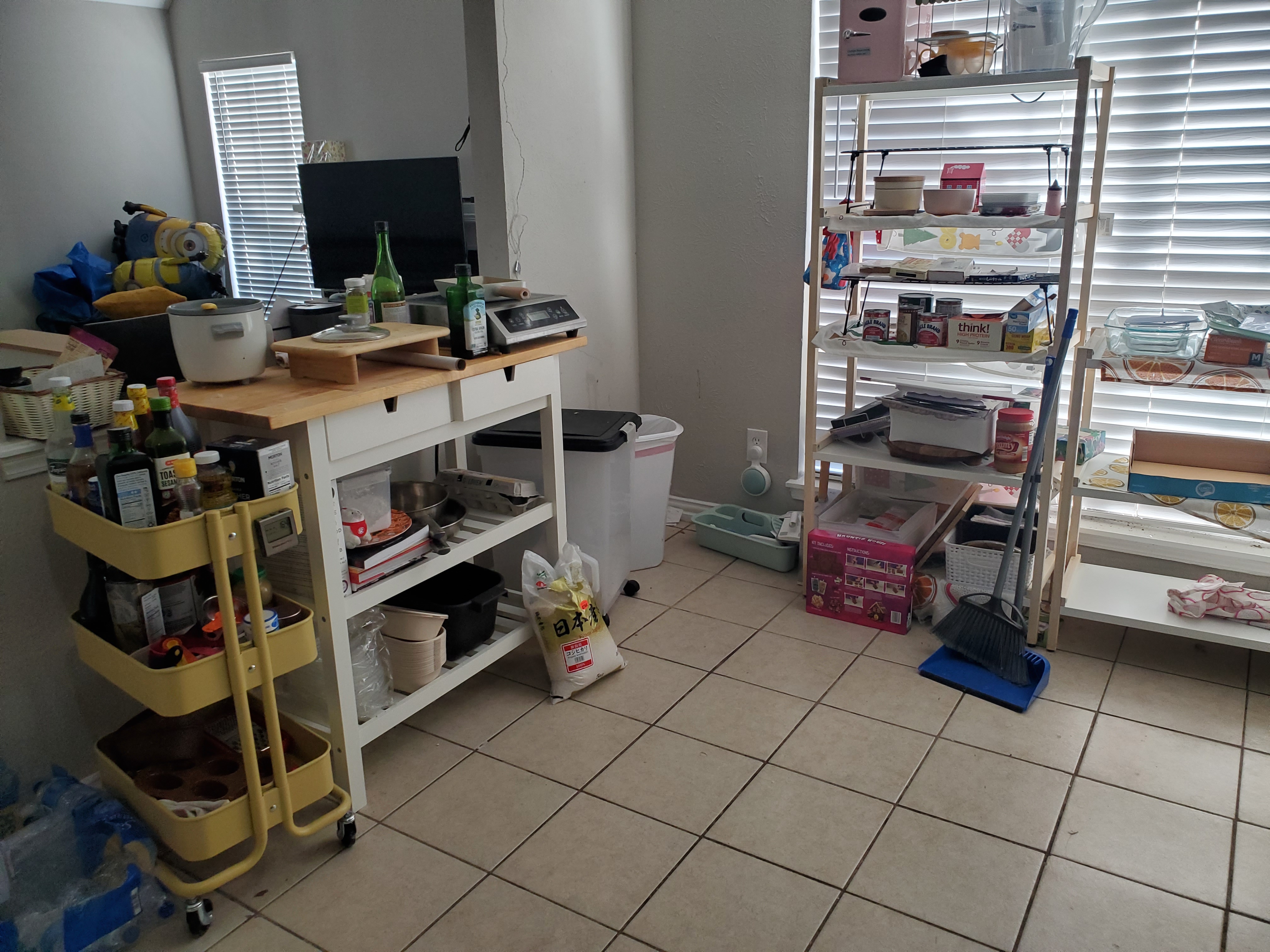 a cluttered kitchen scene with shelves stacked with various items, including food and containers. a yellow cart and a white kitchen island are also filled with kitchenware. the room has tiled floors and blinds on the windows.