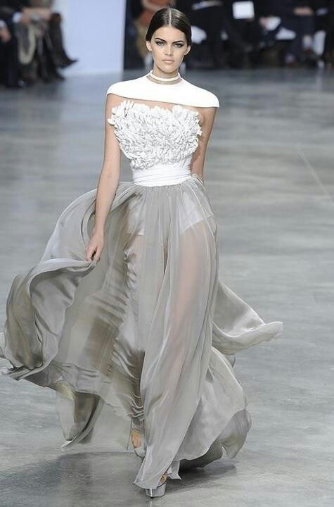 a fashion model walks the runway in a beautiful, white and grey dress. her hair is pulled back, revealing her stunning makeup. she is wearing silver shoes and accessorizing with simple jewelry. the model is striking a pose as she struts down the catwalk.