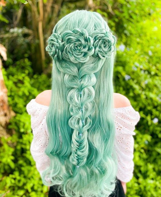 the image shows the back of a woman with long, turquoise hair styled in a unique floral braid. the hair is braided into a crown of roselike shapes at the top, with a complex braid running down the center of the back. the woman is wearing an offshoulder white top. the background is a blur of green foliage.