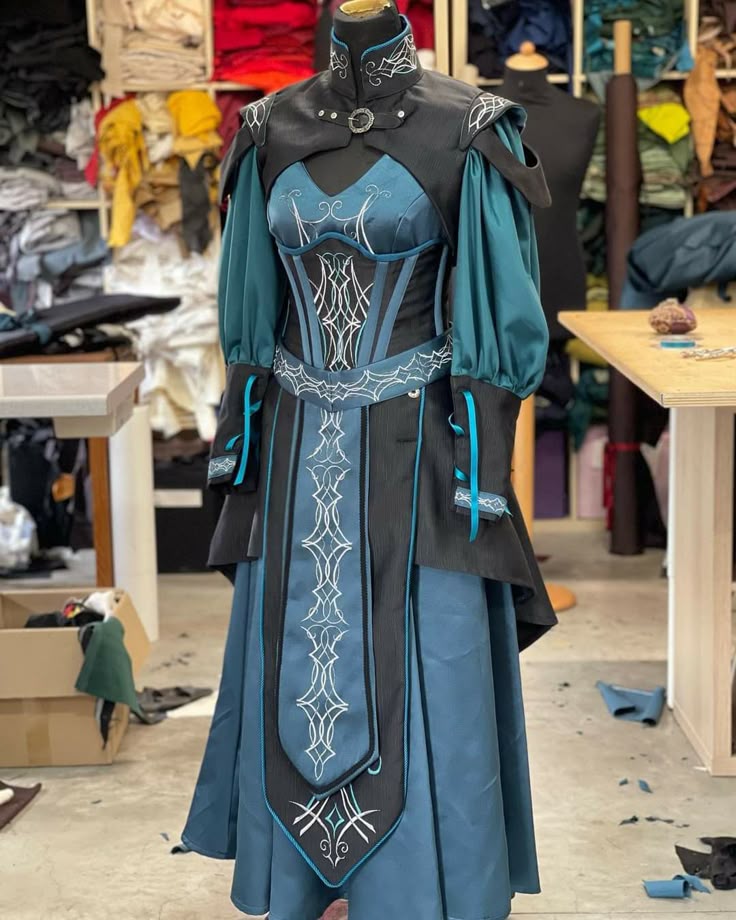 a full view of a teal and black fantasy costume on a mannequin in an atelier. the costume includes a long teal skirt, a teal and black corset with silver embellishments, a black short cape with decorative shoulder pieces, and teal puffy sleeves. the atelier is cluttered with fabrics and sewing equipment.