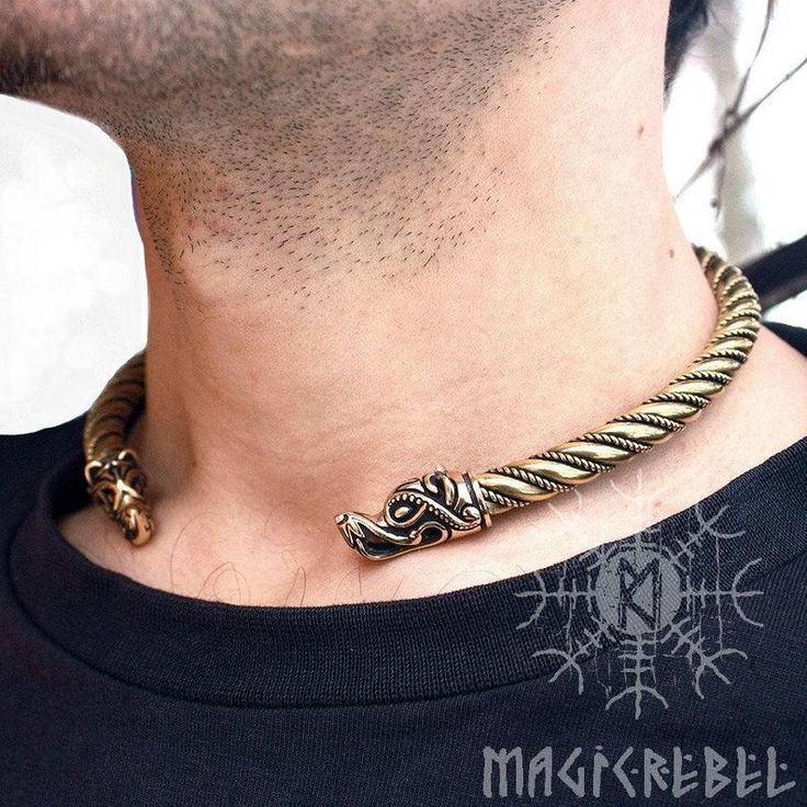 a man is wearing a bronze viking dragon torc necklace. he has a short beard and is wearing a black shirt. the necklace has dragon heads on each end. the background is blurred, but appears to be a room. there is a logo with nordic symbols and the text "magikrebel" overlaid on the image.
