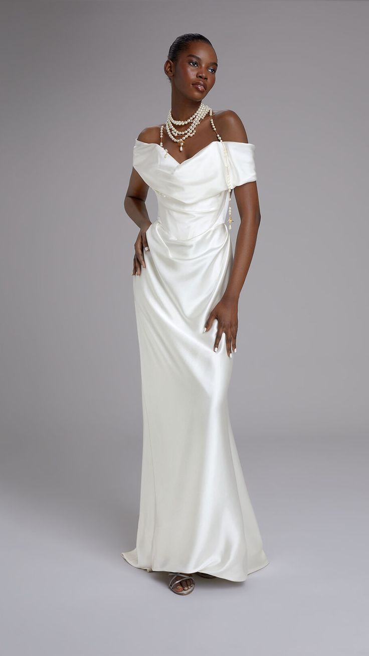 a fulllength studio shot of a darkskinned woman model wearing a simple, elegant ivory offtheshoulder satin wedding dress. she is accessorized with a layered pearl necklace and stands against a gray background. the dress features a draped bodice and a flowing skirt. the overall style is minimalistic and sophisticated.