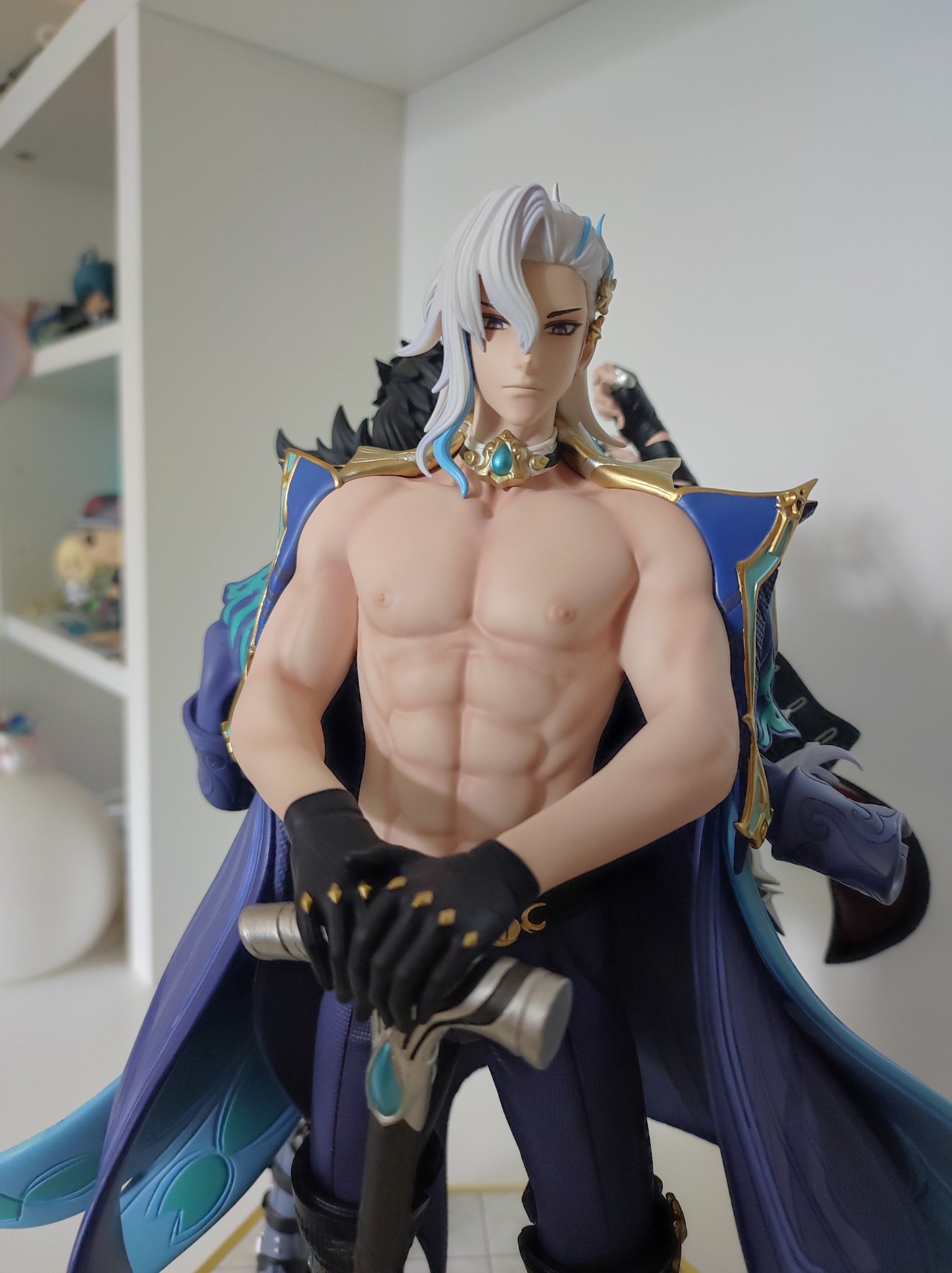 a close up of a male anime figure in a blue and purple outfit with white hair. the figure is holding a silver and blue sword. the figure has a muscular physique. the background is a white shelf with other figures.