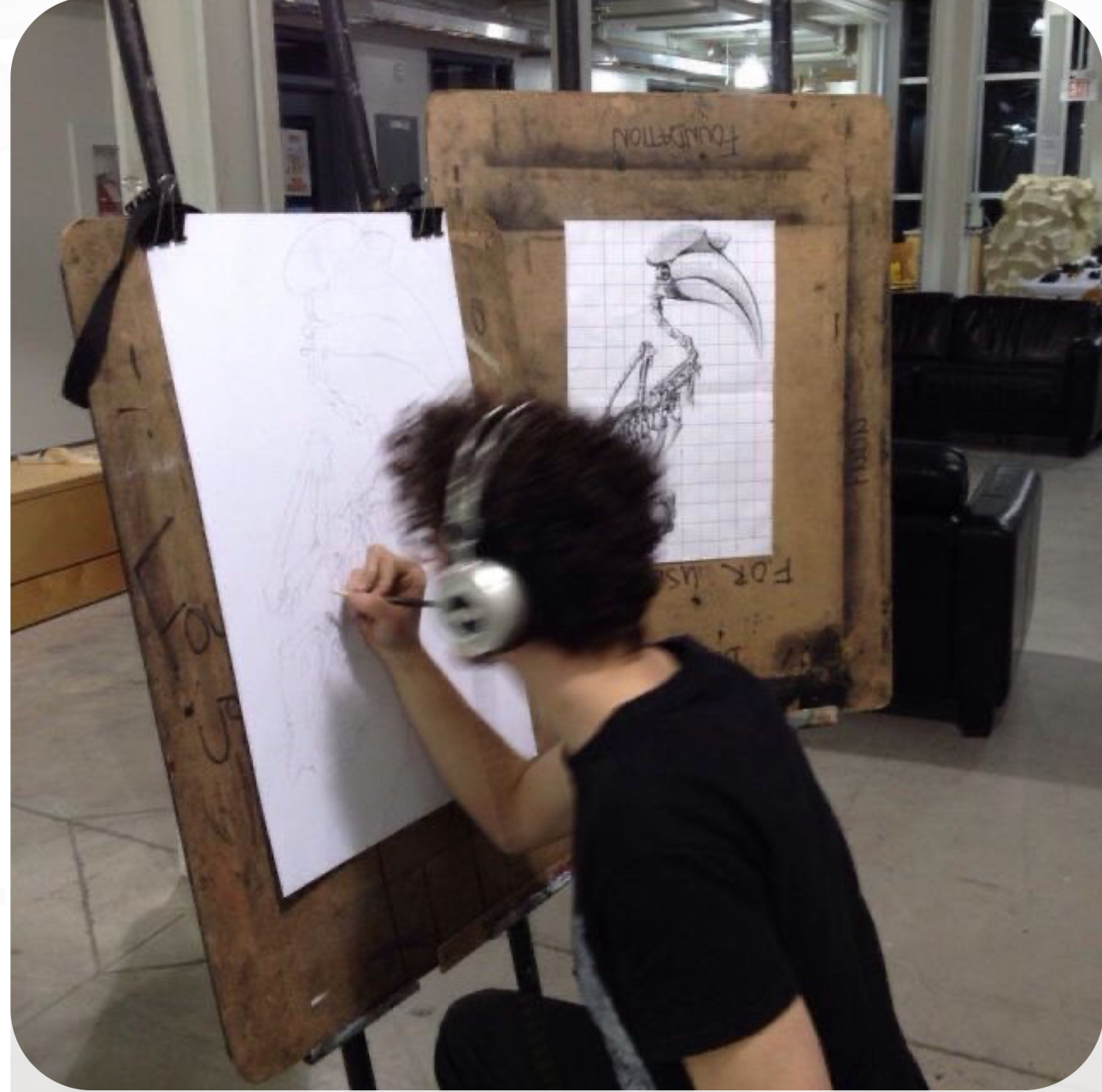 a young man with curly hair is intensely focused on his drawing. he's wearing headphones and is positioned in an art studio, likely a classroom setting, working on a detailed sketch of a bird skeleton. two easels are set up – one displays a gridded reference image, and the other holds his work in progress. the overhead lighting casts soft shadows, emphasizing his concentration.