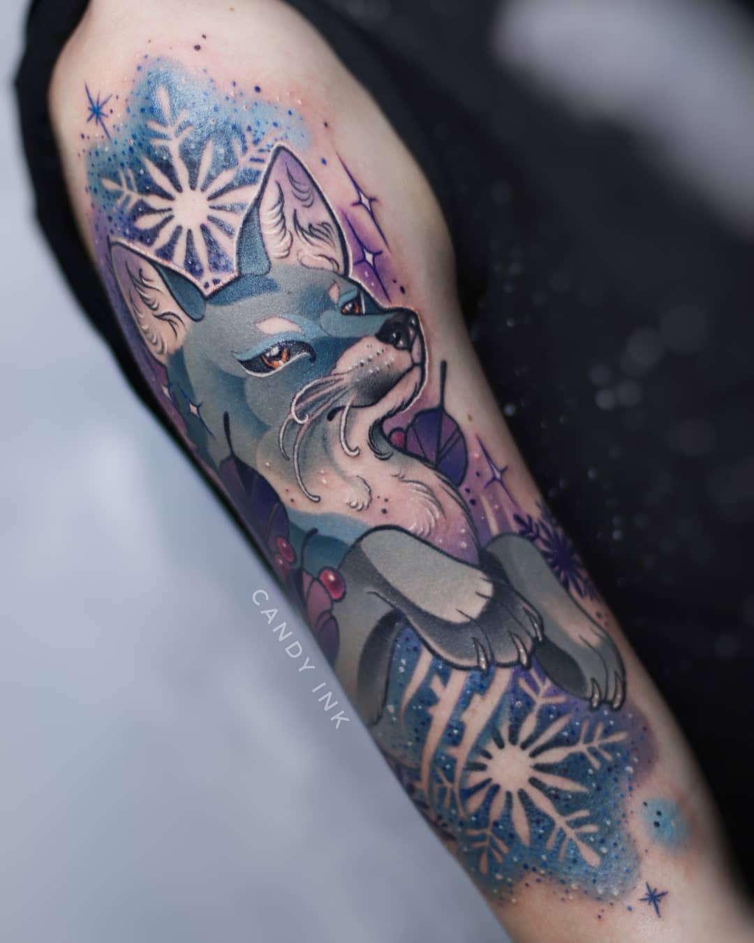 a colorful tattoo of a fox sitting down on someone's upper arm. the fox is surrounded by white snowflakes with a blue watercolor effect as the background. the fox has blue eyes and white fur, with blue shading on its body.  there is a signature reading "candy ink" in white on the bottom left of the tattoo.
