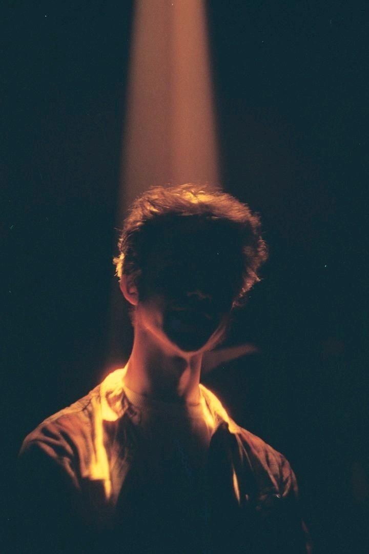 a man with dark hair stands in the shadows with a spotlight shining on his chest, the light illuminating his jacket. the man's face is obscured by the shadows, and a sense of mystery is present. the image has a vintage film aesthetic with grain and a dark, moody feel.