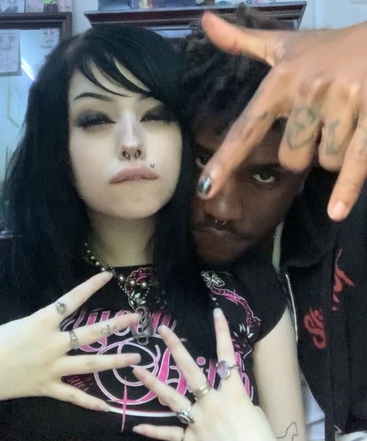 a closeup of a young couple, a blackhaired girl with goth style makeup and a blackhaired boy with tattoos. the couple is close, with the boy's arm around the girl's shoulder. the girl is wearing a black and pink top with a silver necklace, rings on her fingers, and piercings. the boy is wearing a black hoodie and a black tshirt with a design. both are smiling, looking happy and in love. the image is filled with gothic and punk style elements.