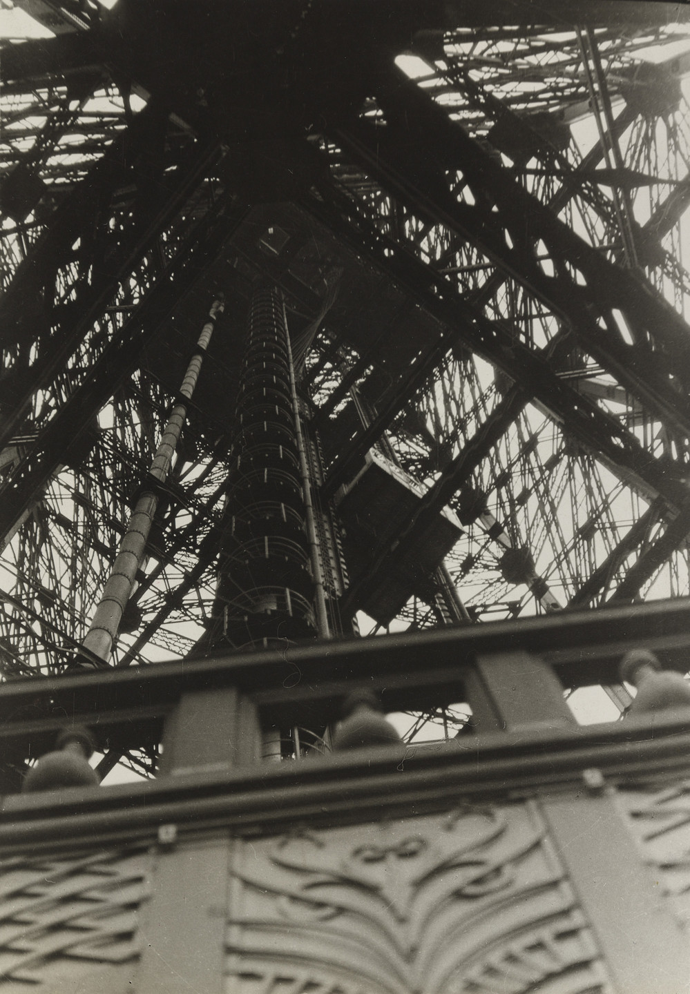 the image is a striking black and white closeup photograph taken from a low angle, looking directly upward into the intricate metal latticework of the eiffel tower. the photograph focuses on the inner structure of the tower, revealing the complex network of beams, columns, and stairs that make up its skeletal frame. a central pillar, containing a staircase and possibly an elevator shaft, dominates the center of the image, drawing the viewer’s eye upwards.  the intricate metalwork is sharply detailed, demonstrating the strength and elegance of the design. in the lower foreground, a section of the tower's ornate decorative railing is visible, providing a contrast in texture and style to the rough, industrial elements of the interior structure.  the overall effect is a powerful image that emphasizes the monumental scale and intricate detail of this iconic landmark.  the perspective and composition create a strong sense of verticality and height, reinforcing the tower's imposing presence.