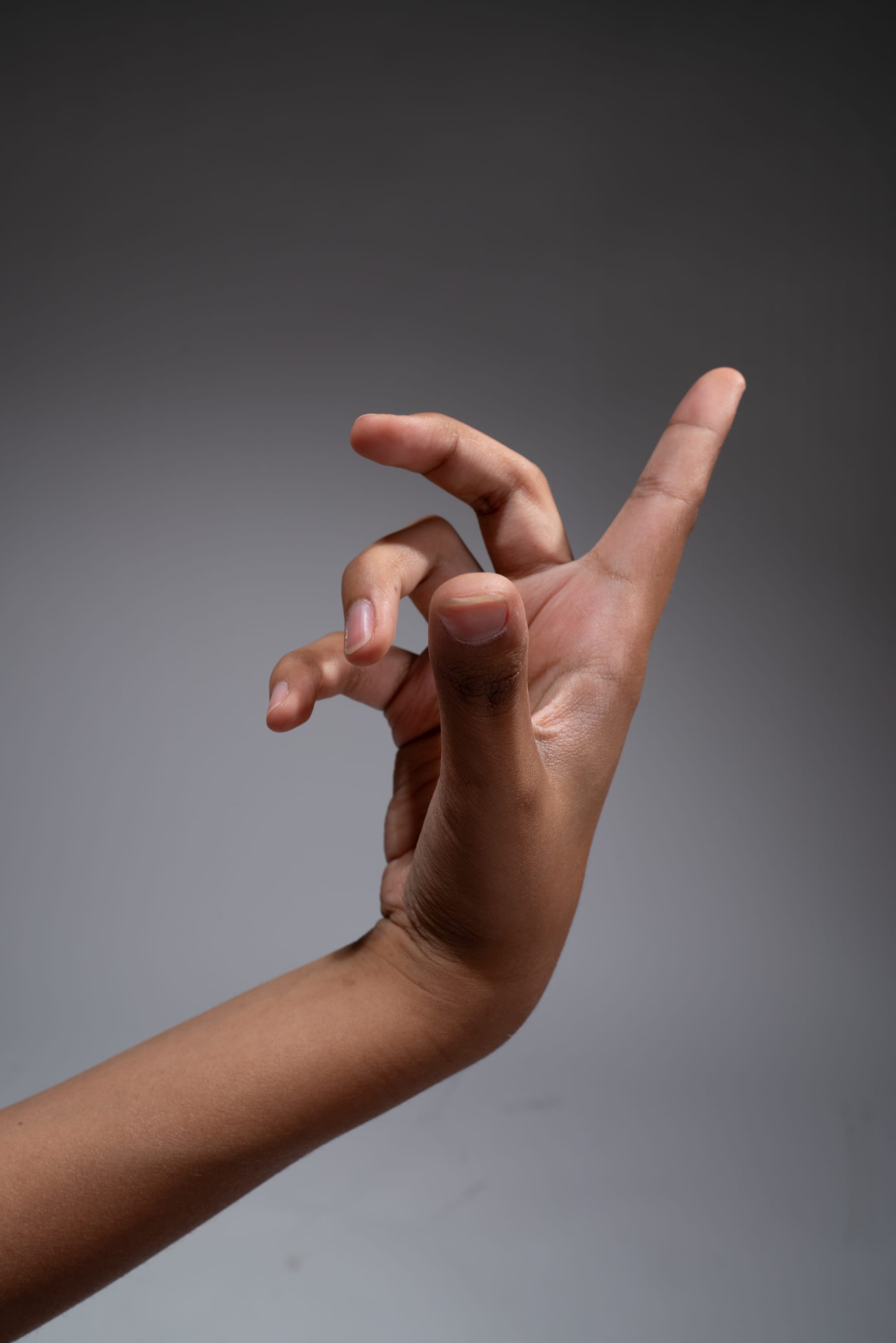 a hand is held up against a gray background. the hand is making a gesture with the index finger pointed up and the other fingers curled inward. the hand is illuminated by studio lighting, which creates a soft shadow on the background.