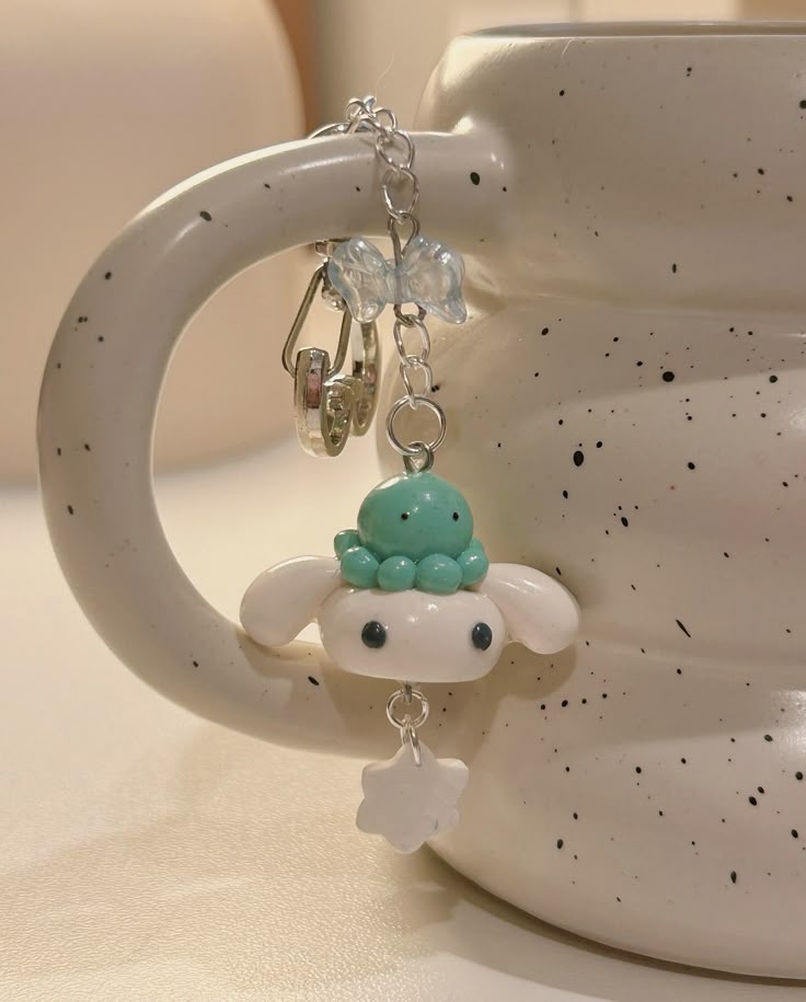 a closeup image shows a cinnamoroll keychain hanging from the handle of a white ceramic mug. the keychain features a white cinnamoroll character wearing a small turquoise octopus on its head. the charm also includes a clear bow, silver chain, and a small white snowflake. the mug is white with black speckles, and the background is a soft, blurred cream color. the image has a bright and cheerful aesthetic.