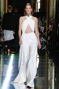 a female model walks the runway in a stunning white jumpsuit with a plunging neckline and a flowing skirt. she is adorned with a delicate necklace and her hair is styled in a sleek updo. the model exudes confidence and elegance, highlighting the sophistication of the design.  she is surrounded by a blurred background of other models and photographers, capturing the energy and excitement of a highfashion runway show. the image showcases the beauty and artistry of contemporary fashion.