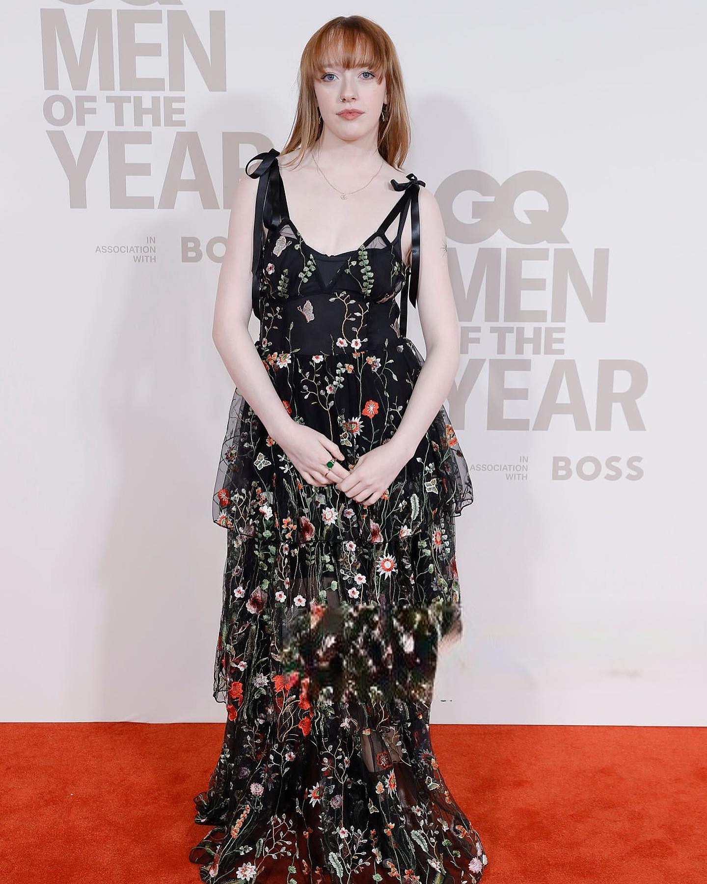 a young woman with red hair, wearing a floorlength black floral embroidered dress with thin straps tied at the shoulders, stands on a red carpet at the gq men of the year awards. the backdrop features the event's branding. she has a calm, poised demeanor, hands clasped gently in front of her. the overall impression is one of elegance and sophistication within the context of a highprofile awards ceremony.