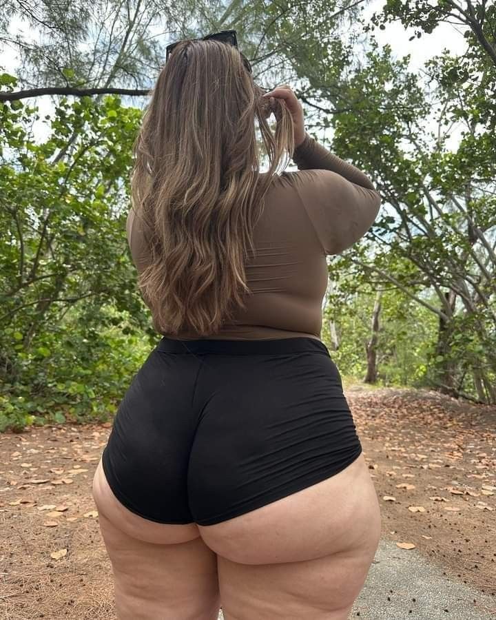 a woman with long brown hair is seen from the back. she is standing on a trail in a forest, wearing a brown longsleeved shirt and black shorts. she has sunglasses on top of her head and is touching her hair. the background is filled with trees and foliage.