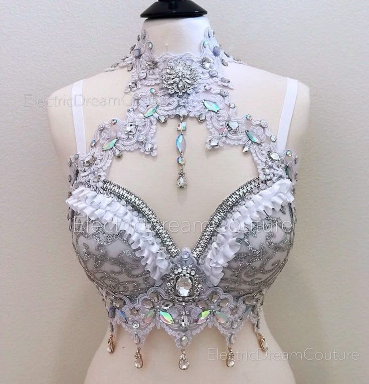 a closeup shot of a bejeweled white and silver bustier or bra on a mannequin. the bustier features intricate lace detailing, ruffles, and a high neckline, embellished with a variety of gemstones and crystals. the design also includes hanging crystal pendants and a central large crystal embellishment. the branding "electricdreamcouture" is visible on the image.
