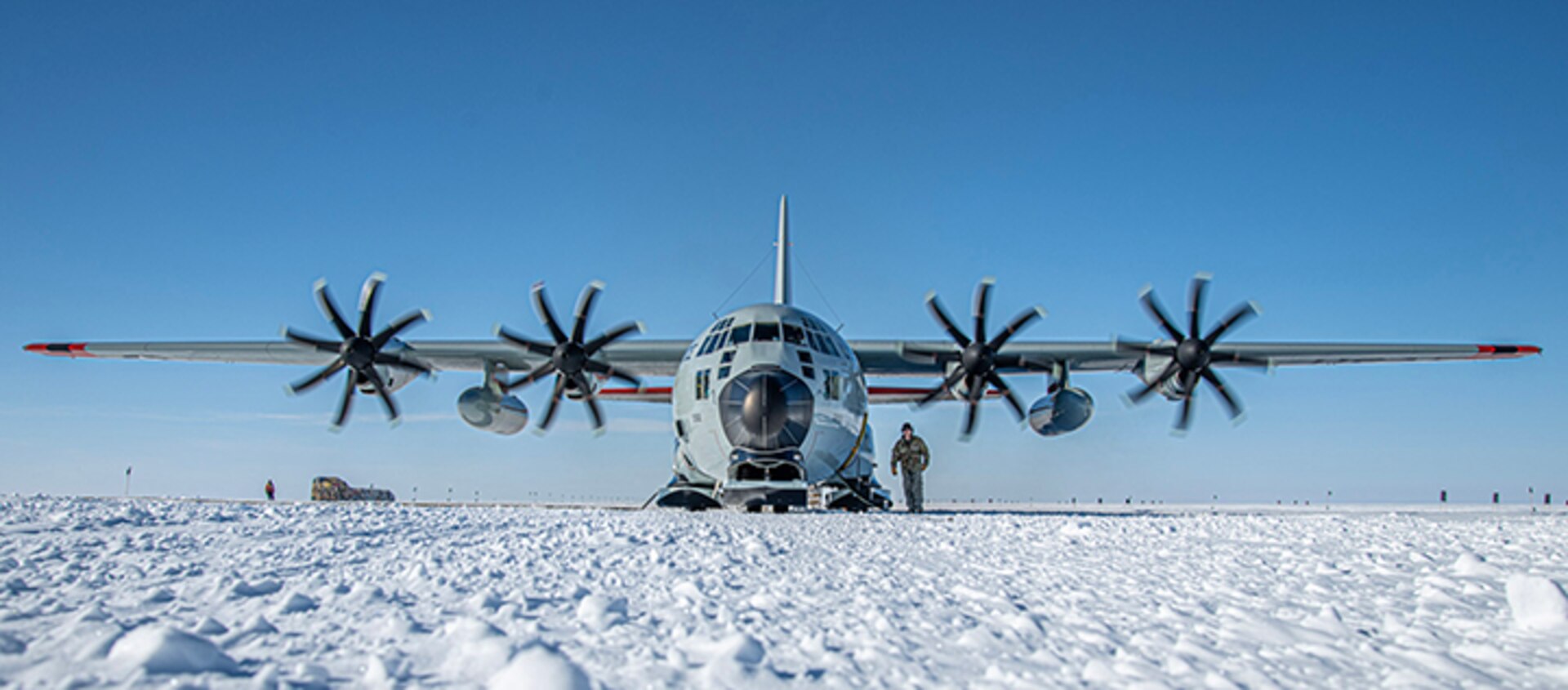 a military transport aircraft, a c130 hercules, is parked on a snowcovered runway in front of a blue sky. the plane is facing the camera, with its propellers visible and spinning. the snow is fresh and white, and there are a few other aircraft in the distance. the plane is being loaded with cargo for a mission.