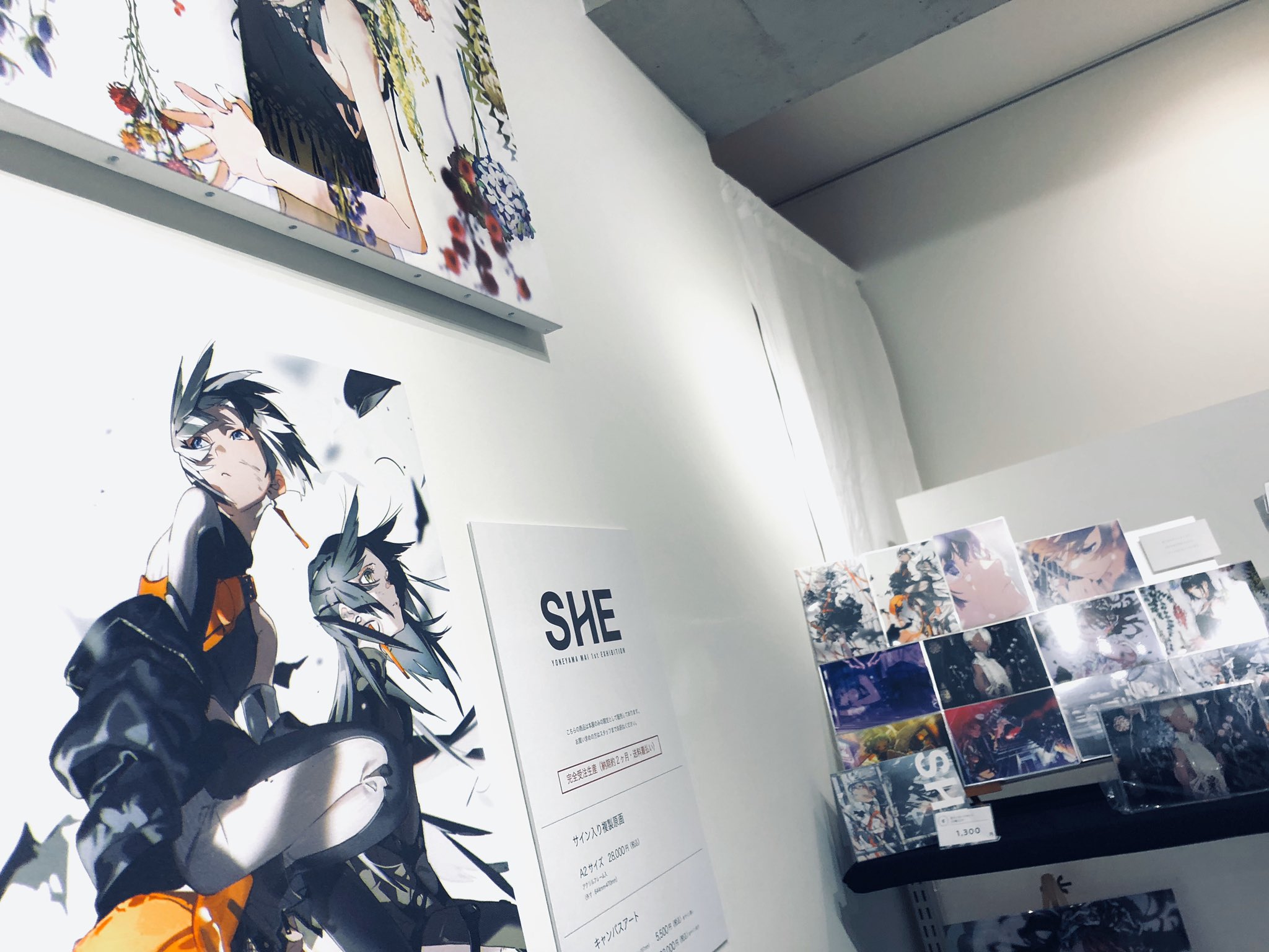 the image shows a corner perspective of an art exhibition featuring animestyle art. the brightly lit space showcases multiple posters and artwork on display, while a shelf to the right likely holds prints and merchandise. the overall aesthetic uses a black and white palette with pops of color.