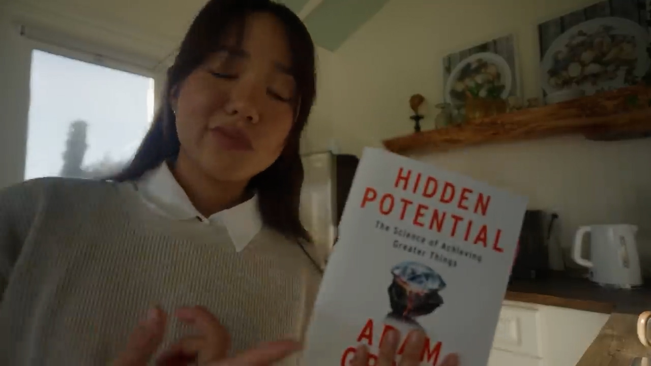 a young woman is standing in a brightly lit kitchen and reading a book titled 'hidden potential'. the image is taken from eye level and shows the woman from the waist up. she is wearing a white sweater and has her hair down. the kitchen is decorated in a minimalist style with white walls and wooden accents.