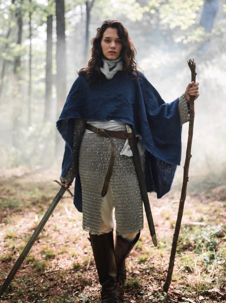 a woman in medieval attire stands in a forest. she wears chainmail, a blue tunic, boots, and a belt. she carries a sword and a staff, and appears to be a warrior or adventurer.