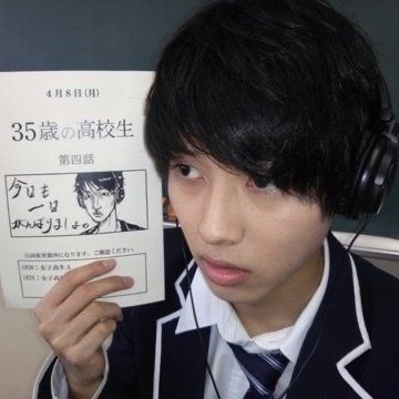 a young asian man is taking a selfie while holding a piece of paper in his hand. the paper appears to be a title card for a japanese drama. the man is wearing a school uniform with a white shirt and a black jacket. he has short, dark hair and brown eyes. the image is brightly lit and the man is looking directly at the camera.