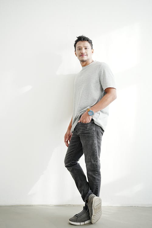 a man is standing against a white wall, wearing a gray tshirt, gray jeans, and gray sneakers. he has a watch on his left wrist and is looking at the camera.