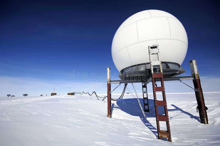 a large, white radar dish stands tall on a snowy, white landscape in antarctica. the radar dish is mounted on four tall, rustcolored metal legs. the background of the image is a bright blue sky.  the image was taken during the day. the view is wide and horizontal.