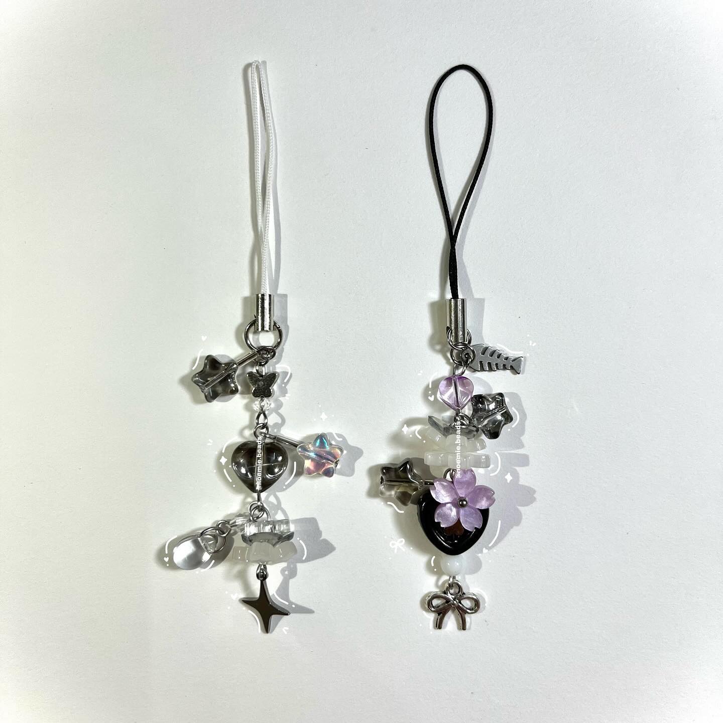 two handcrafted phone charms are displayed against a white background. the charm on the left has a white strap and a collection of silver charms including a star, butterfly, heart, and a fourpointed star. it also has some iridescent beads. the charm on the right has a black strap and a similar collection of charms in silver, including a fish, star, heart, flower, and bow. it also has purple beads.