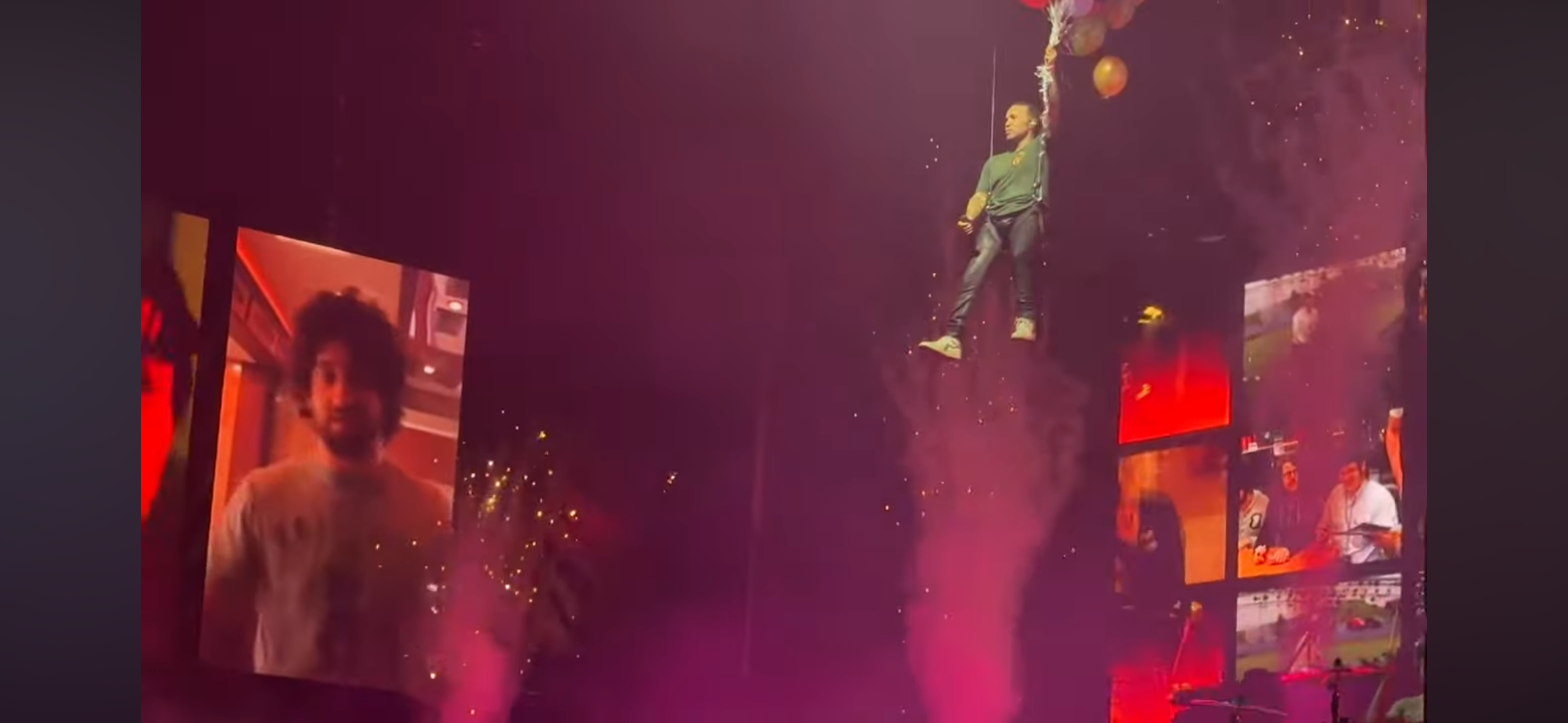 a concert performance with a singer suspended in the air by balloons. the stage is dimly lit and there are screens on either side of the stage displaying footage of the performer. there are also lights shining down on the stage. the audience is cheering and waving their hands in the air.
