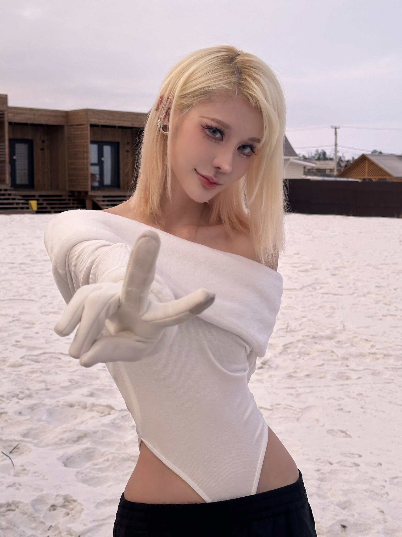 a young blonde woman wearing a white offtheshoulder bodysuit and white gloves poses outdoors in the snow. she's making a peace sign with her hands. in the background, there are some houses. the overall aesthetic is stylish and elegant.