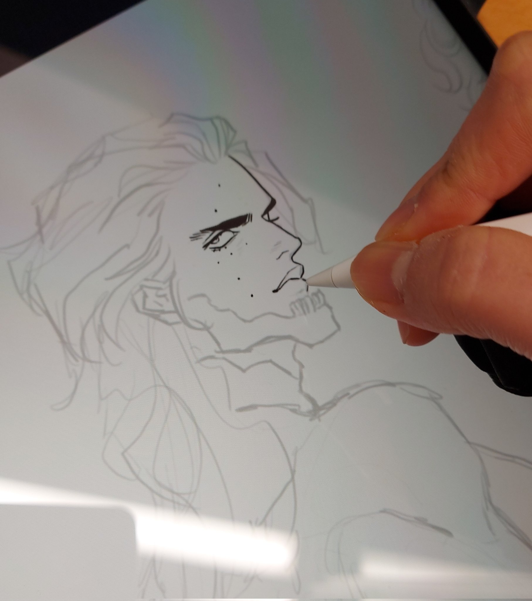 a closeup image shows a hand holding a digital pen and drawing a sketch on a tablet. the sketch depicts a male figure with detailed facial features and long hair, with an element of a skeleton incorporated into the jawline. the style suggests a digital art illustration in progress.