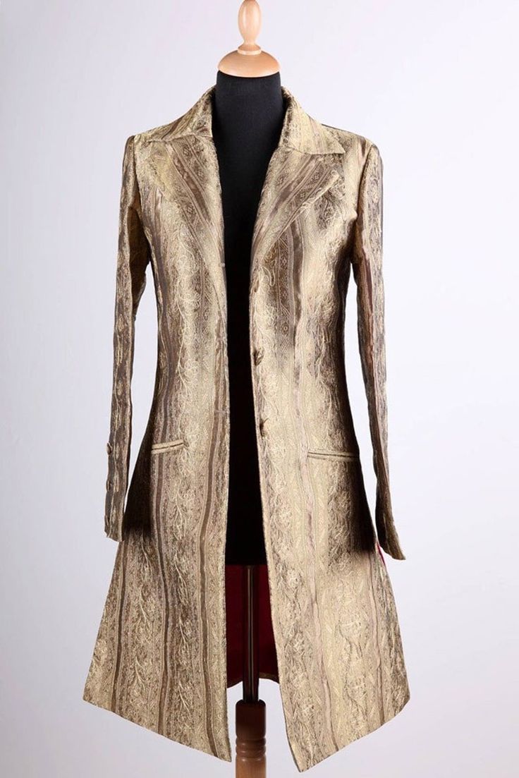 a closeup shot of a gold brocade women's coat displayed on a mannequin against a white background. the coat is a long, fitted style with a collar, lapels, and button closures. it features a decorative brocade design and a red lining. the coat is an example of vintage fashion.