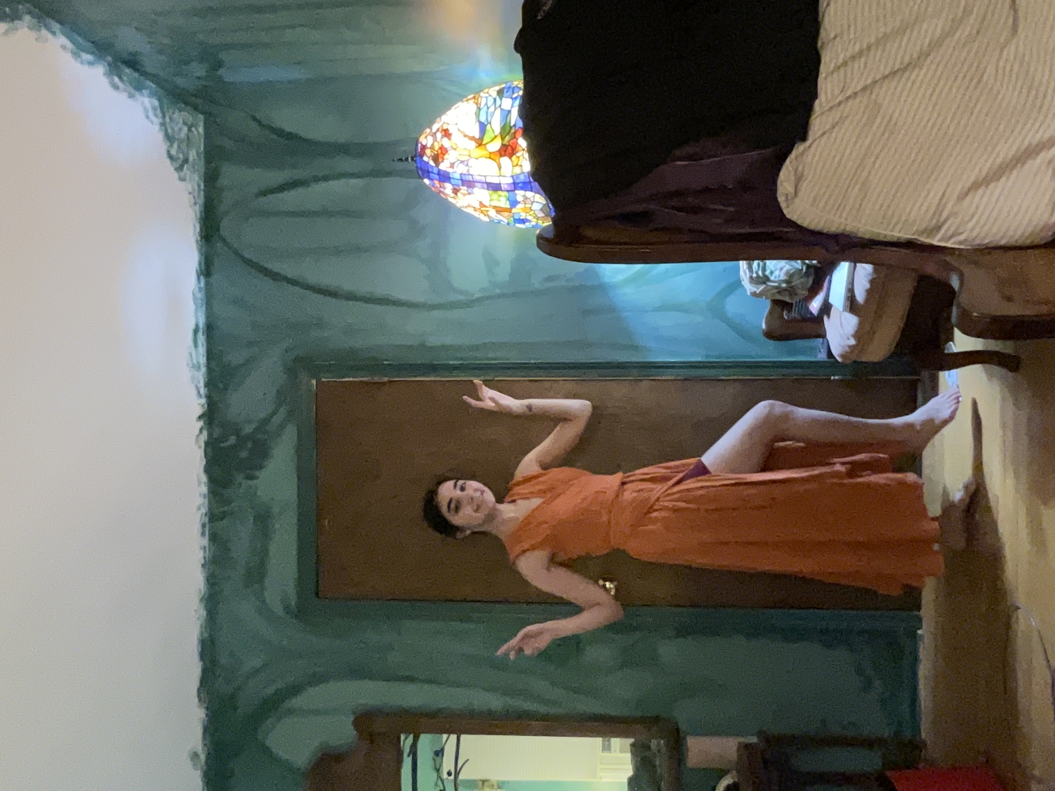 a young woman in a long, orange wrap dress playfully poses in a bedroom with teal painted walls. the walls have a textured, almost marbled effect. she's positioned in front of a door and partially in front of an armchair and bed. a stained glass lamp is visible in the background, adding to the room's eclectic atmosphere.  the overall feel is casual and relaxed.