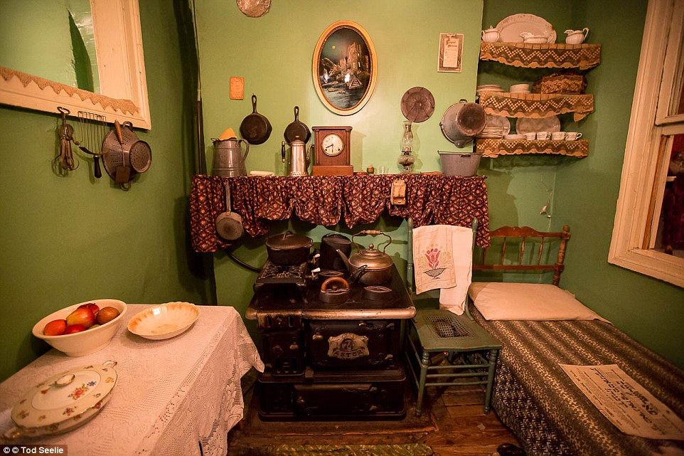 the image showcases a meticulously preserved historical kitchen, likely from the late 19th or early 20th century. the room is small and sparsely furnished, painted in muted green tones. a woodburning stove dominates the center, flanked by a narrow bed on the right and a small table set for a meal on the left.  shelves laden with dishes and cookware line the walls, while vintage utensils hang from hooks. the overall effect is one of humble simplicity, offering a glimpse into the domestic life of a bygone era.