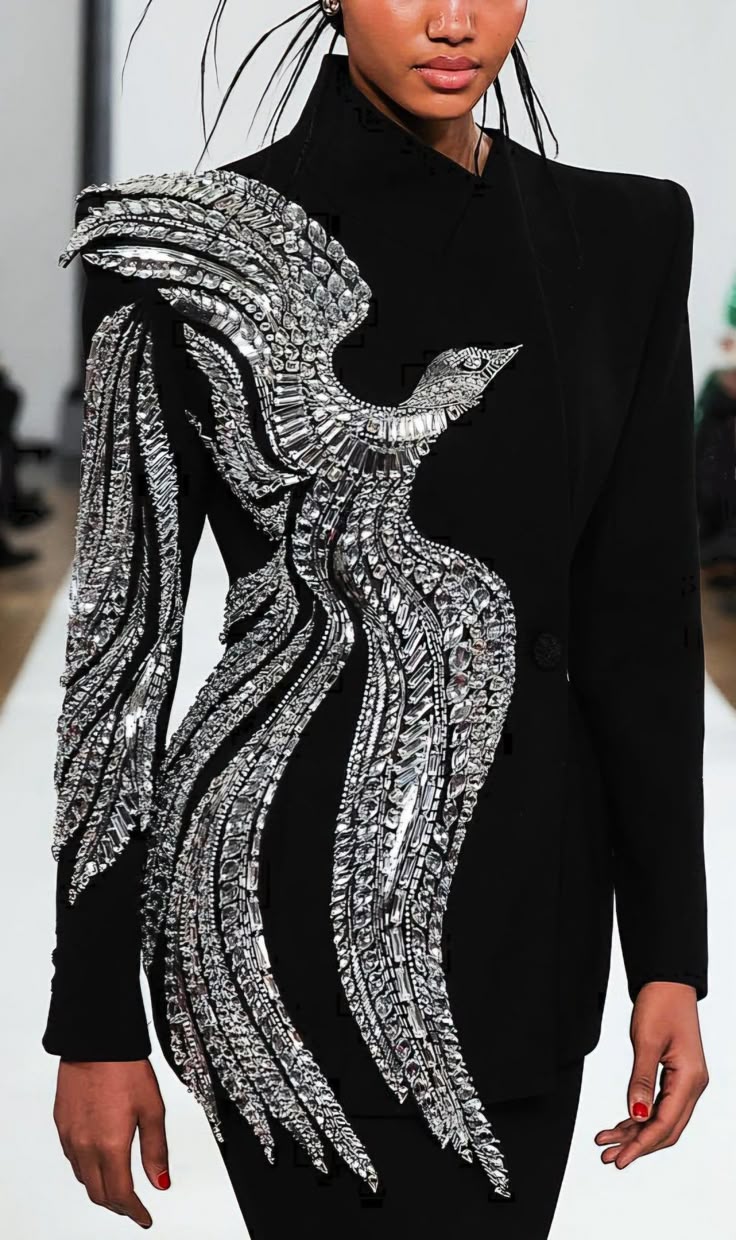 a model wearing a black dress with a large, ornate silver bird design on the bodice is seen on a runway. the bird design is made of silver embellishments and extends from the shoulder down the side of the dress. the model has dark skin and is wearing subtle makeup. she is standing in front of a blurry background.