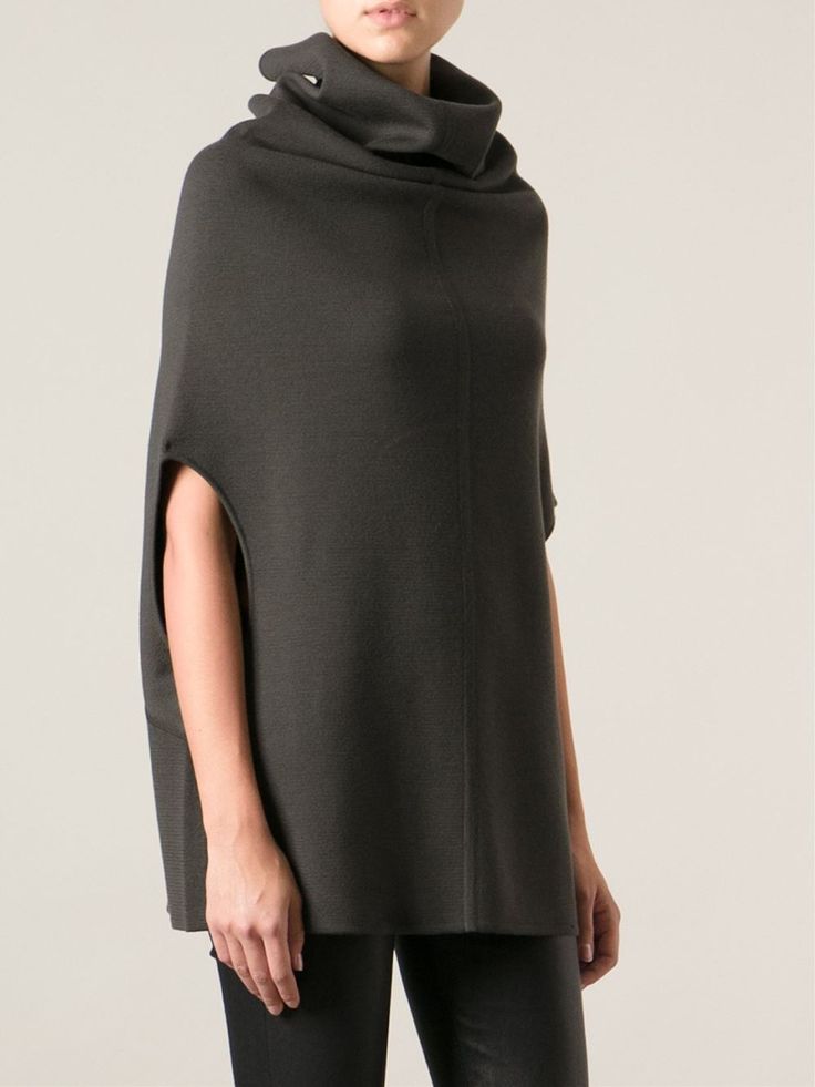 a woman models a dark gray cowl neck poncho. the poncho has an opening for her arms. she is wearing black pants or leggings. the background is a light neutral color.