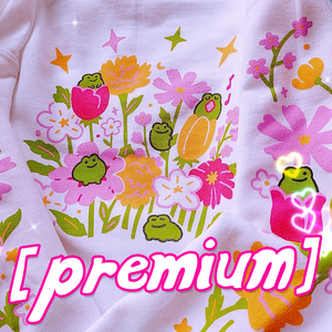a closeup shot of a white sweatshirt featuring a whimsical design of pink, yellow, and orange flowers with cute green frogs nestled amongst them. the overall aesthetic is pastel, cute, and kawaii.  a large pink "premium" label is overlaid on the image. the sweatshirt appears soft and comfortable.