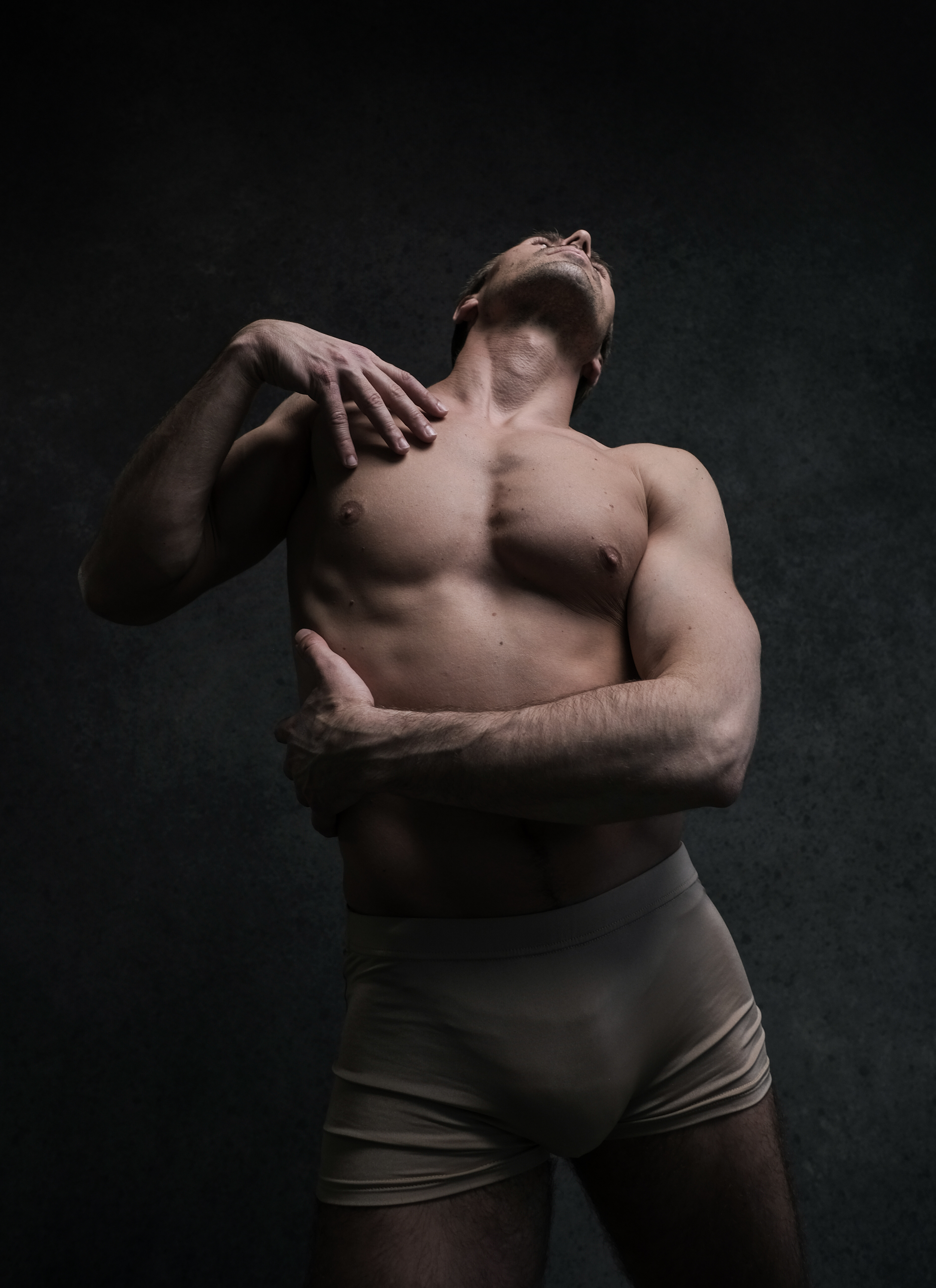 a muscular man in nude color underwear is posing in a dance studio. his body is lit dramatically, showcasing his physique. the background is dark and textured.