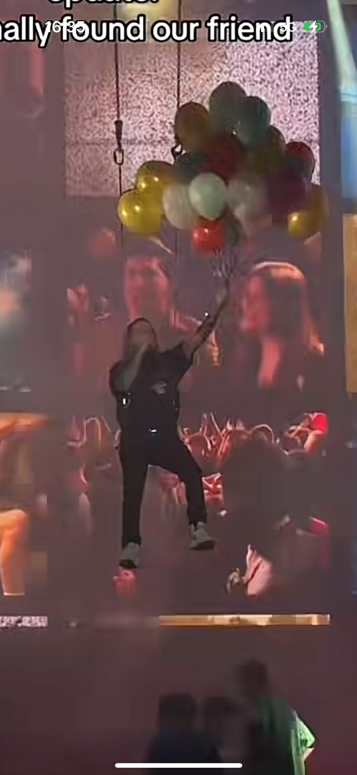 a person on stage is singing into a microphone while holding a bunch of balloons. the crowd is cheering and waving their hands in the air, with a big smile on their faces. the stage is lit up with bright lights and the atmosphere is electric. it seems like they are happy to have finally found their friend.
