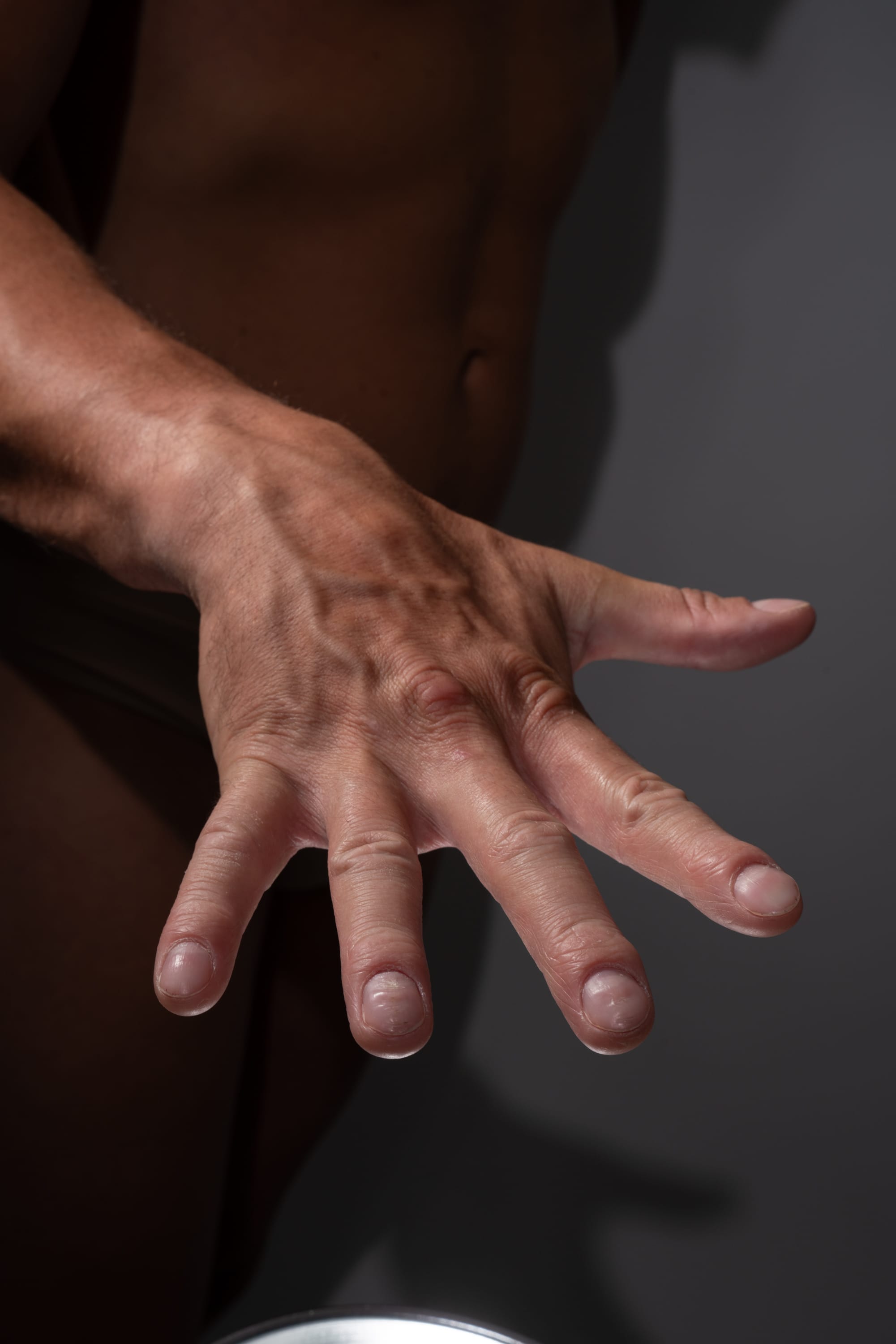 the image shows a closeup of a man's hand reaching out towards the viewer. the hand is muscular with prominent veins and tendons visible beneath the skin. the lighting is soft and highlights the texture and detail of the hand.  the overall color palette is warm and muted.