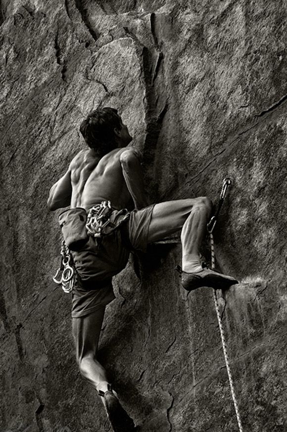 a black and white image of a male rock climber ascending a steep rock face. the climber is wearing a harness and has a rope attached to his waist, along with other climbing gear. he is focused on the next hold as he reaches up with his left hand and continues to climb the crag. the image captures the intensity and focus required for this challenging sport.
