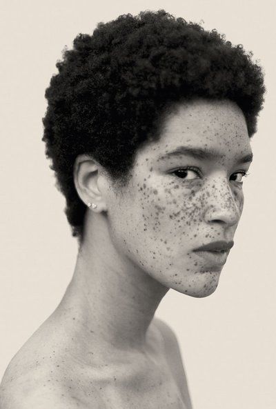 a black and white portrait of a woman with freckles and short, curly black hair. she is looking to the side, and her face and neck are visible. she is wearing small earrings.