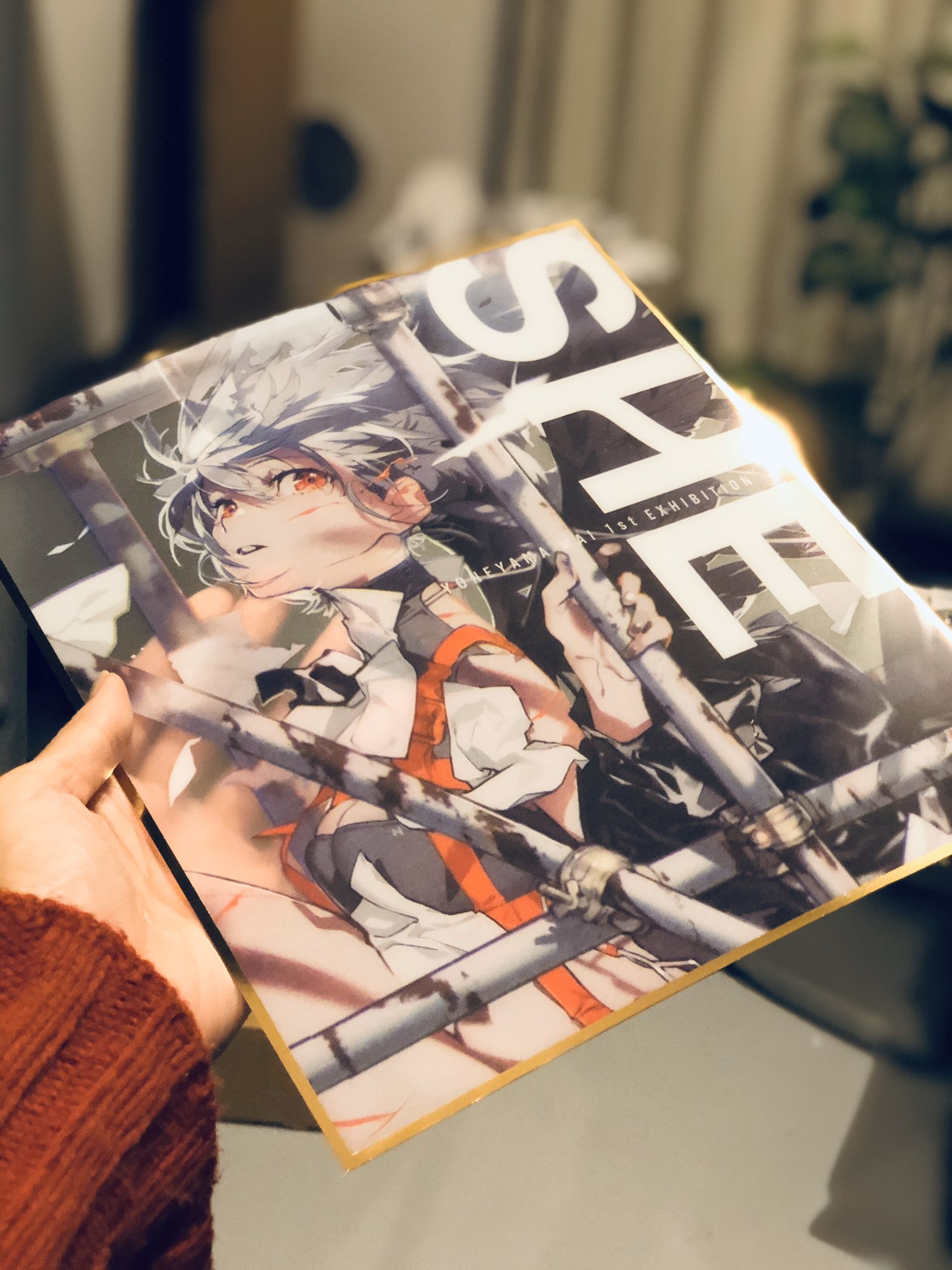a person is holding up an anime art print featuring a female character with white hair and a black and red outfit. the background of the print is abstract and urbanthemed. the print seems to have a gold foil finish. the photo is taken indoors with warm lighting.