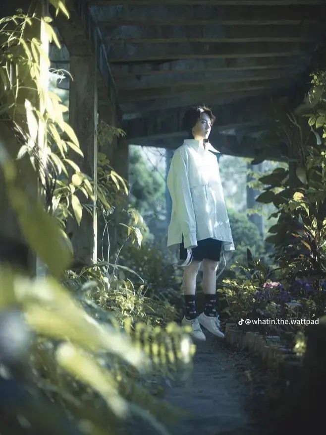 a young adult with short hair wearing a white oversized shirt, black shorts, and white sneakers stands in an overgrown garden. they are framed by a crumbling concrete structure, with lush greenery surrounding them on all sides. the photo is taken at eye level and uses natural, diffused light, creating a soft and serene atmosphere.