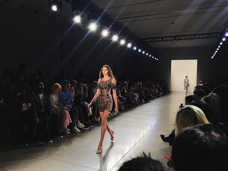 a fashion show is in progress with a model walking down the runway wearing a plaid dress and heels. an audience is seated on both sides of the runway watching the show. bright lights illuminate the runway and the space is indoors.