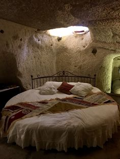 a cozy bedroom inside a cave is shown, with a round bed prominently featured. the bed is dressed in white bedding with a ruffled skirt and is adorned with multiple throw pillows and a patterned throw. a wrought iron headboard sits at the top of the bed. above, a skylight allows natural light to enter the cave, illuminating the unique space. the walls and ceiling of the cave are rough and textured, adding to the room's rustic and intimate ambiance.