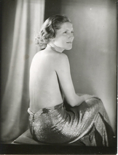 the photograph is a black and white portrait of a woman from the 1930s. she is seated and partially nude, wearing a shimmering, metalliclooking skirt that pools around her.  her back is mostly exposed, and she's turned slightly away from the camera, looking over her shoulder. her hair is styled in a classic 1930s wave. the background features a draped curtain, which creates a soft, diffused light.  the overall mood is one of elegance and glamour, characteristic of art deco photography. the image has a timeless quality, suggesting a studio portrait from a bygone era.