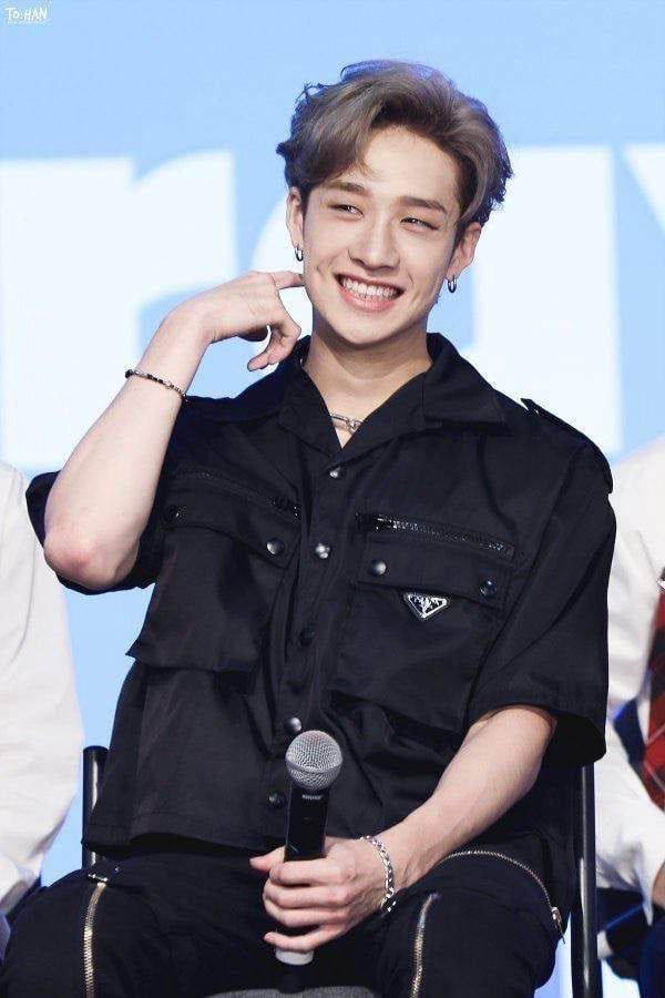 a closeup shot of bang chan, a member of the kpop group stray kids, smiling brightly. he's wearing a stylish black prada shirt, silver bracelets, and a necklace. his hair is styled in a silvergrey look.  he's holding a microphone, suggesting an interview or fansign event. the background is a light blue. the overall impression is one of friendly approachability and youthful energy.